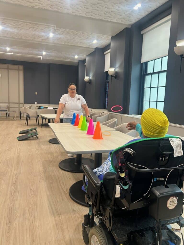 Caregiver standing at a table with colorful cones arranged in a row for a game, as a person in a motorized wheelchair watches in a bright room with windows.