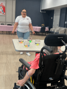 Smiling woman in a white shirt leads a game at a table with fish bowls; an elderly person in a wheelchair watches nearby.