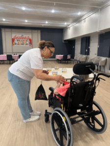 Caregiver leaning toward an elderly person in a wheelchair, holding hands in a bright event room with a carnival banner in the background.