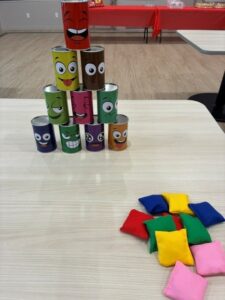 Pyramid of decorated tin cans with cartoon faces stacked on a table; colorful fabric cushions lie nearby on the right side.