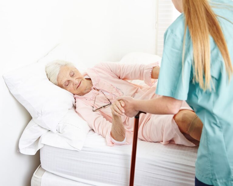Elderly woman in pink pajamas lying in bed, receiving assistance from a caregiver in teal scrubs who is helping her stand with a cane nearby.